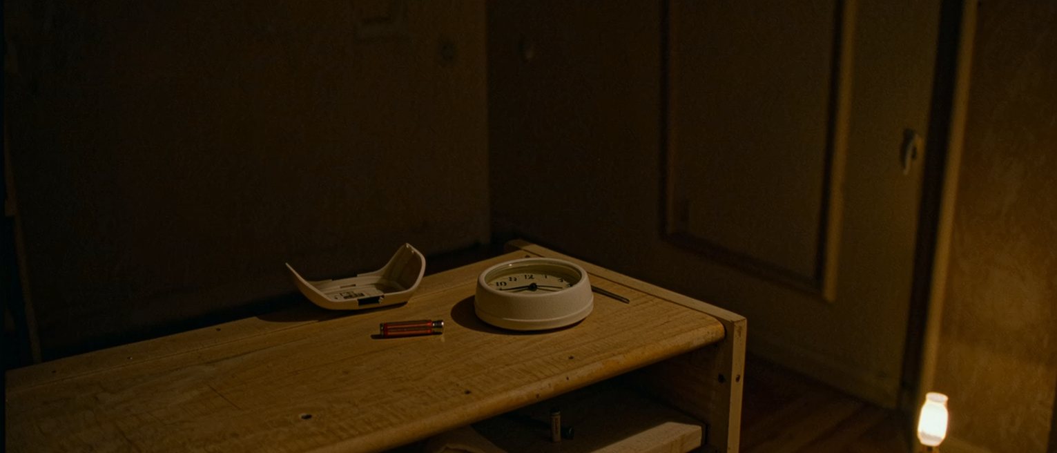 a cupboard shelf seen close with a round white alarm resting with its cover lifted off beside a spare battery and a small screwdriver, a folded cloth, the shelf boards soft beyond