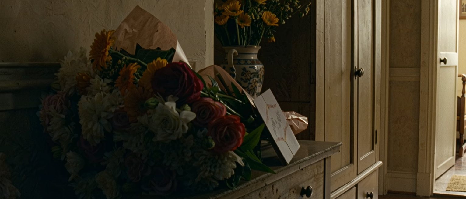 a hall table with a bunch of flowers left in their wrapping beside an unopened card, the vase put back in the cupboard