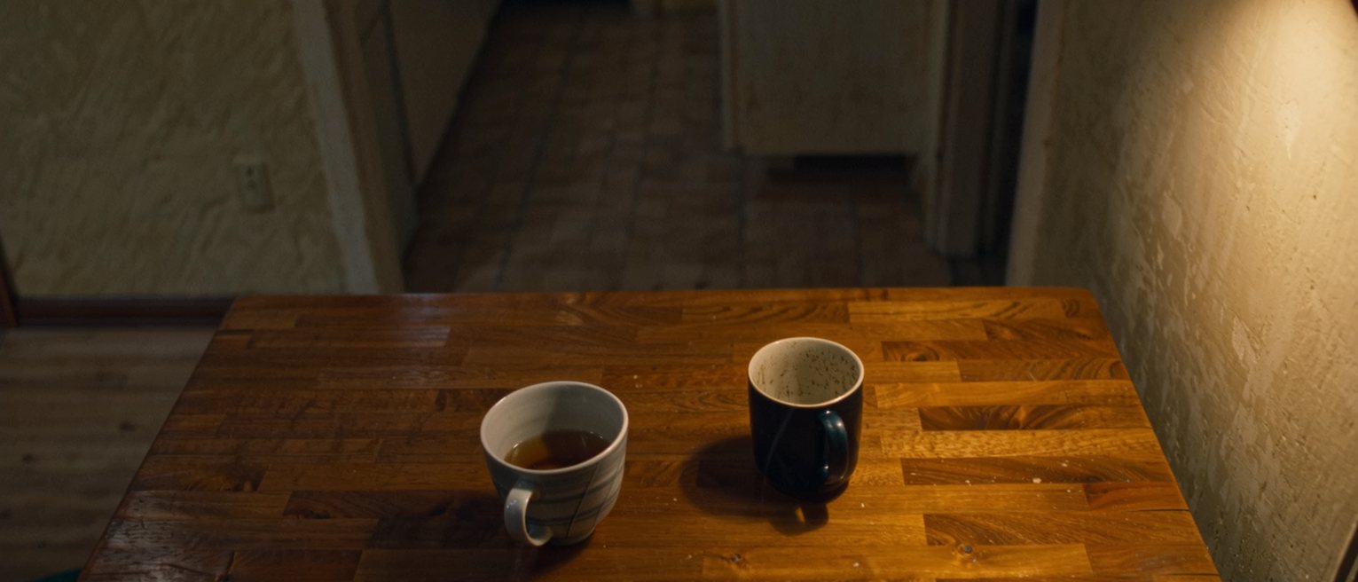 a kitchen table with a second cup set out and no one coming, the tea gone cold in the pot