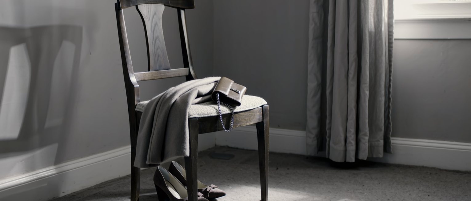 a bedroom chair seen close with a coat folded over it, a pair of good shoes set together beneath, and on the seat a small clutch bag with its clasp open, the curtains soft behind