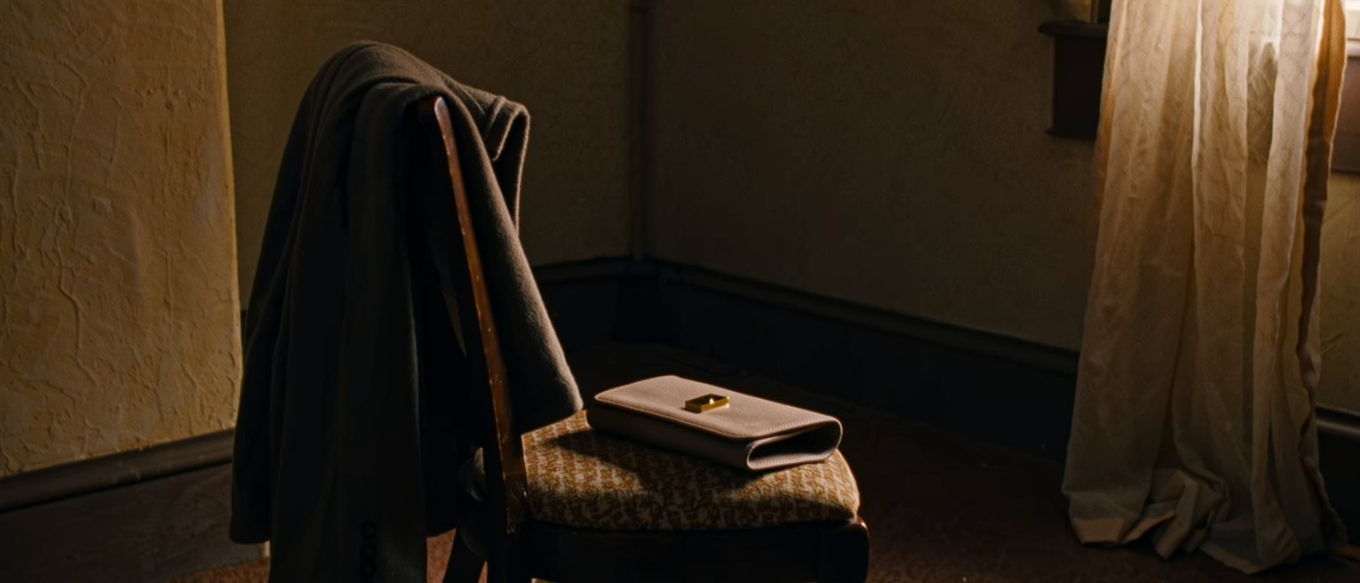 a bedroom chair seen close with a coat folded over it, a pair of good shoes set together beneath, and on the seat a small clutch bag with its clasp open, the curtains soft behind