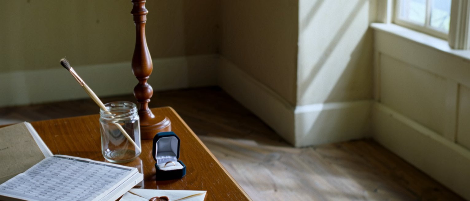 a drawing room seen close with a sketchbook open beside a ledger of household accounts, a paintbrush in a jar, a ring box open and empty on the table and a letter with a wax seal, the lamp close behind