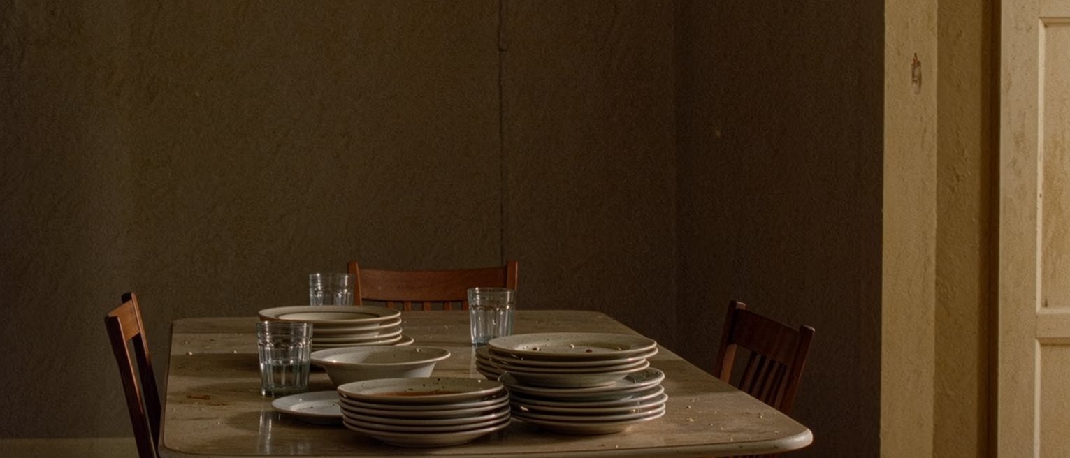 a kitchen table seen close after a supper for eight, every plate used and stacked high, glasses everywhere, a bowl of fruit and a dresser full of china close behind, and one chair at the near corner turned a little away from the rest