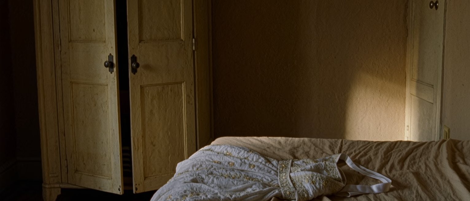 a bedroom with a dressing gown laid across the made bed and a pair of slippers set beneath it, the wardrobe door standing open and the window soft behind