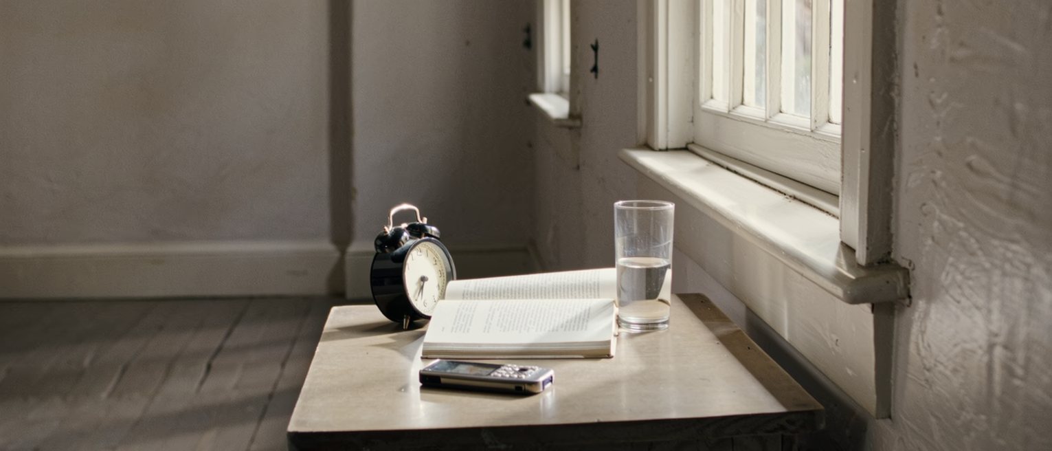 a bedside table with a phone face down, a half-read novel and an untouched glass of water, the alarm clock turned to face the wall