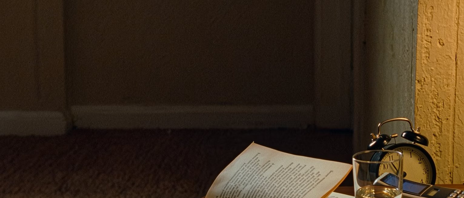 a bedside table with a phone face down, a half-read novel and an untouched glass of water, the alarm clock turned to face the wall