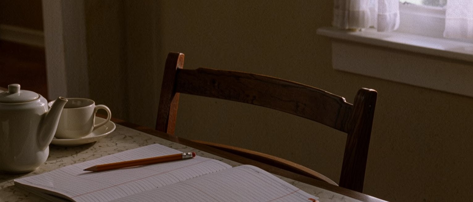a dining table seen close with an open notebook ruled into two columns in pencil, a pen laid across it, a teapot and one cup, the second chair pushed in on the far side, the window soft beyond