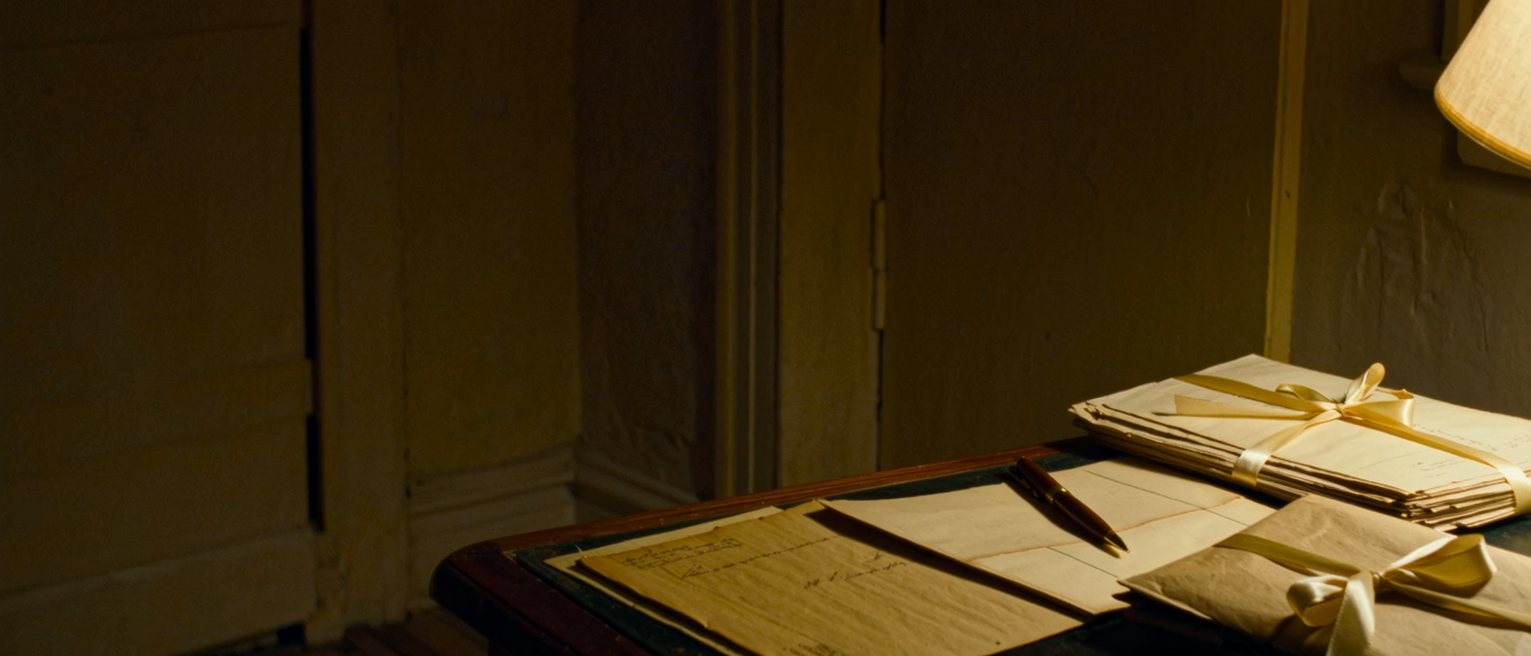 a desk with a bundle of old letters tied with ribbon beside a single new one opened flat, a pen set down across it