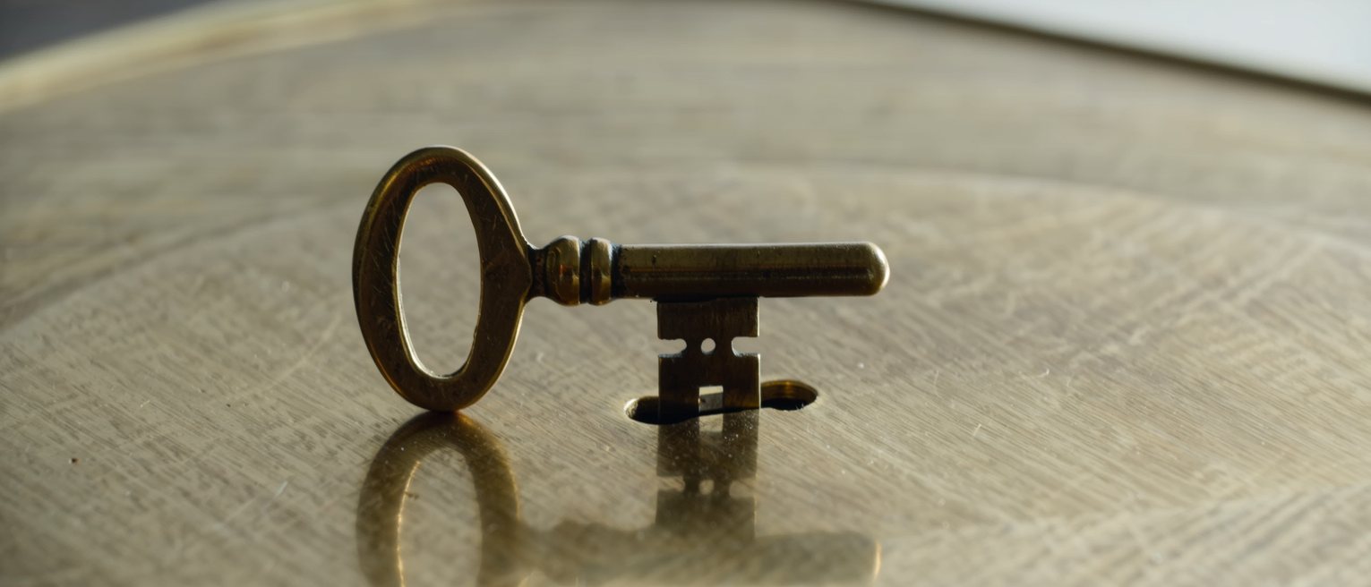 a flat brass clock winding key with an oval bow handle, standing upright with its shaft seated in the winding hole of a brass clock plate