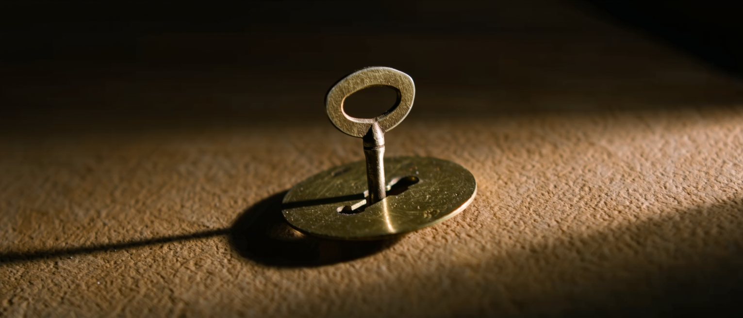 a flat brass clock winding key with an oval bow handle, standing upright with its shaft seated in the winding hole of a brass clock plate