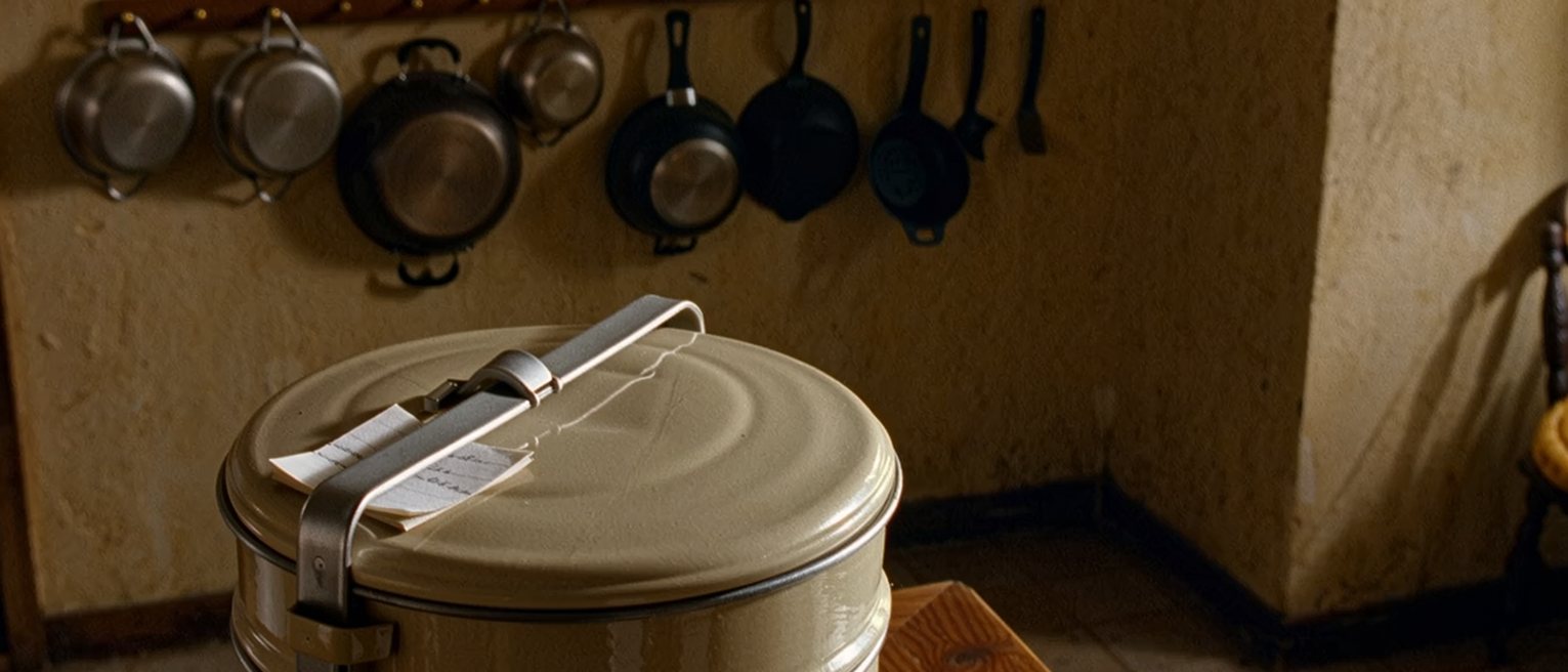 a kitchen counter with a stacked tiffin carrier clipped shut and a folded note tucked under its clip, the cooking things washed and racked behind