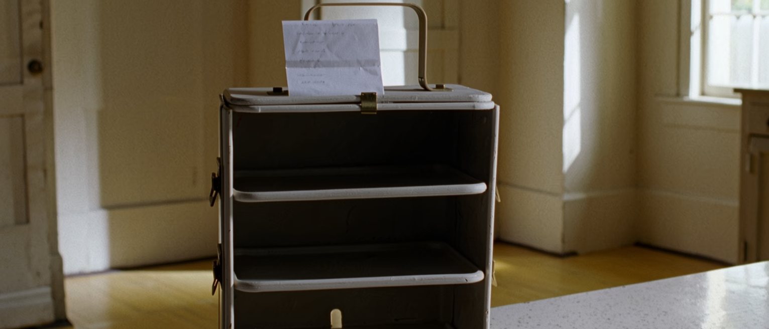 a kitchen table with a tiffin carrier opened into its three tiers, every one of them emptied, and a folded note propped against the top tier