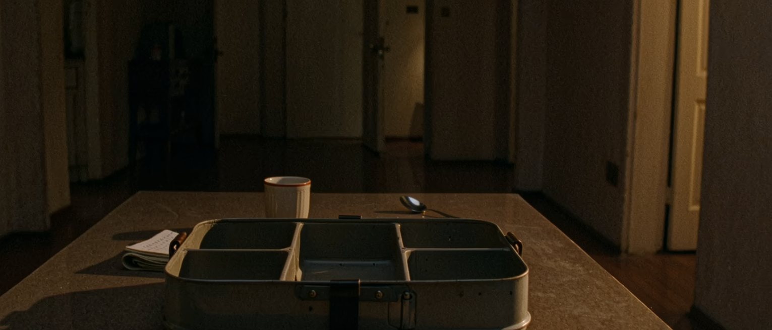 a kitchen table with a tiffin carrier opened into its three tiers, every one of them emptied, and a folded note propped against the top tier