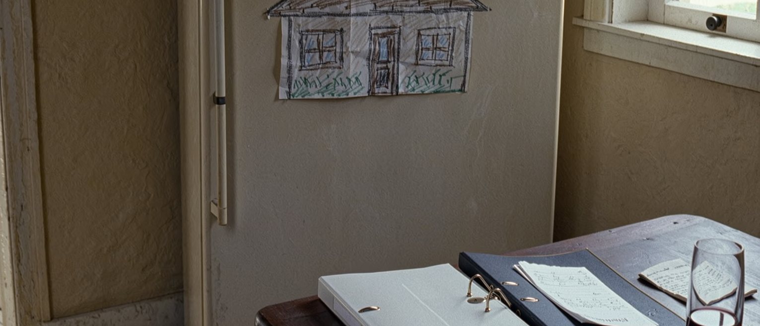a kitchen table seen close with two folders from two law firms, a handwritten list folded small and a glass of wine, a crayon drawing of a house pinned to the fridge close behind