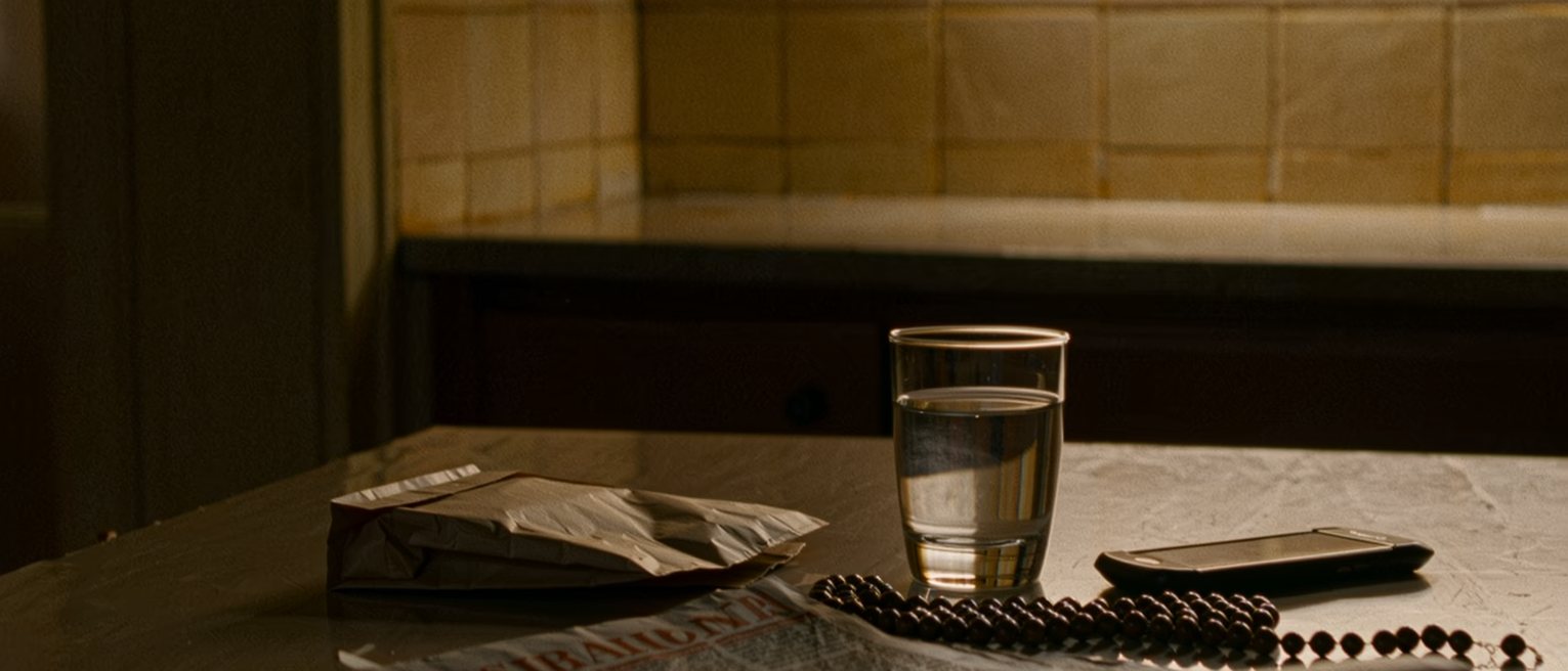 a kitchen table seen close with a rosary coiled beside a folded newspaper, a glass of water, a pharmacy bag and a mobile phone face down, the tiled wall soft behind