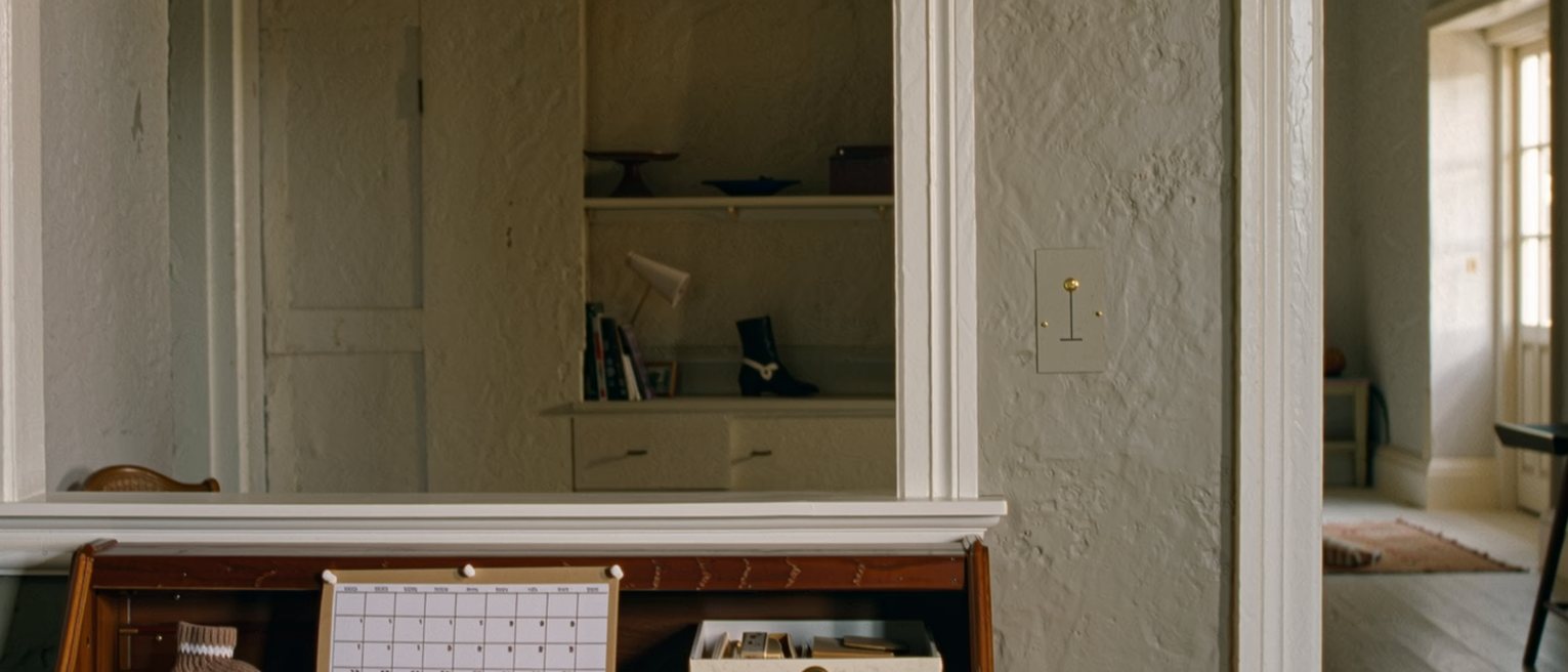 a desk seen close with a wall planner pinned with nothing written in the far months, a calendar of appointments beside it and a small knitted bootee in a box, the shelves soft behind