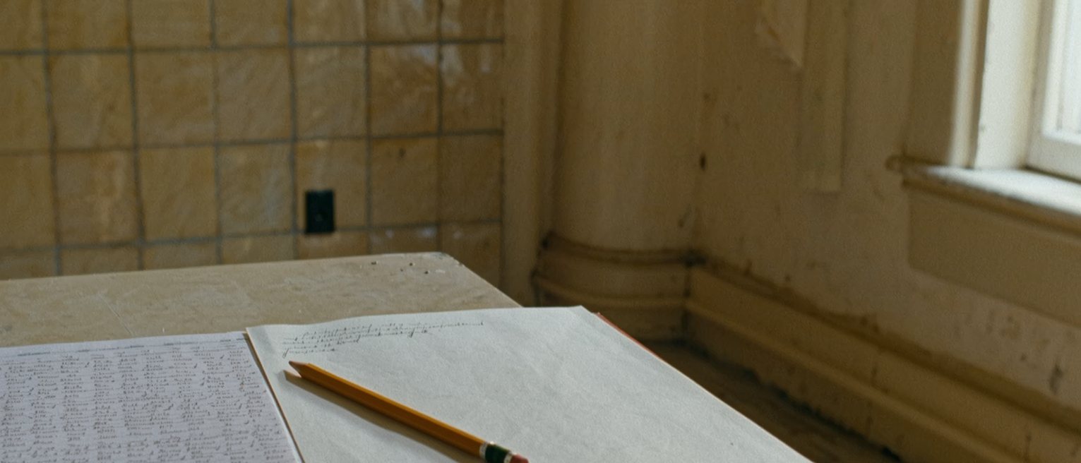 a kitchen table seen close with two exercise books, one filled edge to edge and the other with a single page written, a shared pencil between them and the tiled wall soft behind