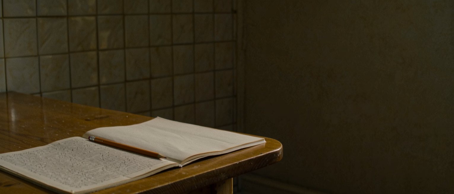 a kitchen table seen close with two exercise books, one filled edge to edge and the other with a single page written, a shared pencil between them and the tiled wall soft behind