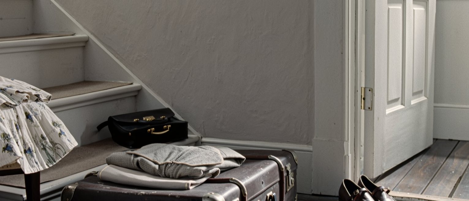 a hall seen close with one suitcase packed and strapped and one pair of shoes left by the door, a folded dress on the chair and a leather satchel on the step above, the stairs soft behind