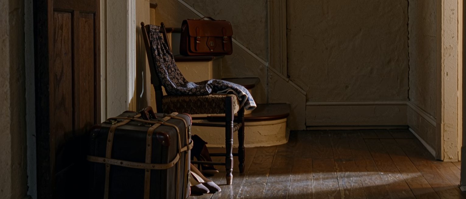 a hall seen close with one suitcase packed and strapped and one pair of shoes left by the door, a folded dress on the chair and a leather satchel on the step above, the stairs soft behind