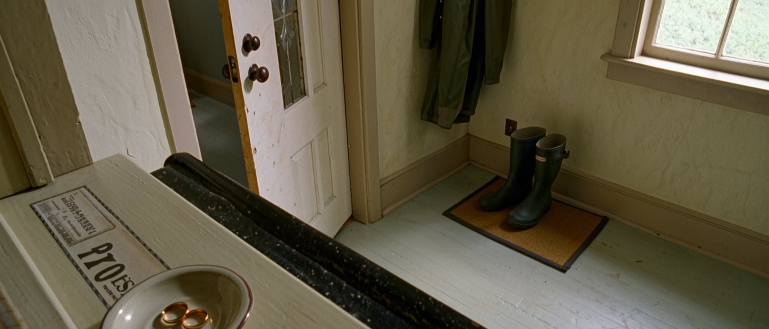 a hall seen close with two plain rings in a dish on the console beside a licence stamped by the registry, a pair of gumboots on the mat and a raincoat on the hook, the door with its glass close behind