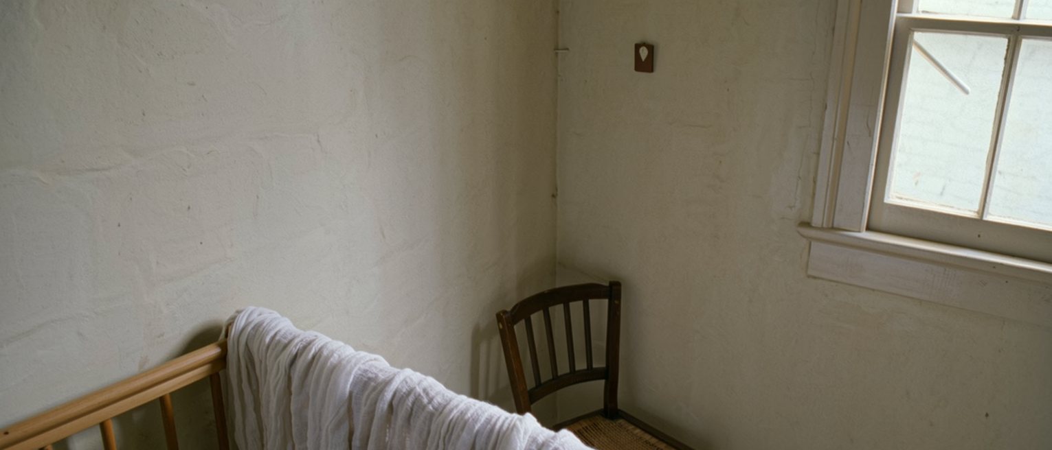 a nursery corner seen close with a cot rail and a folded muslin hung over it, a small chair pushed against the wall behind