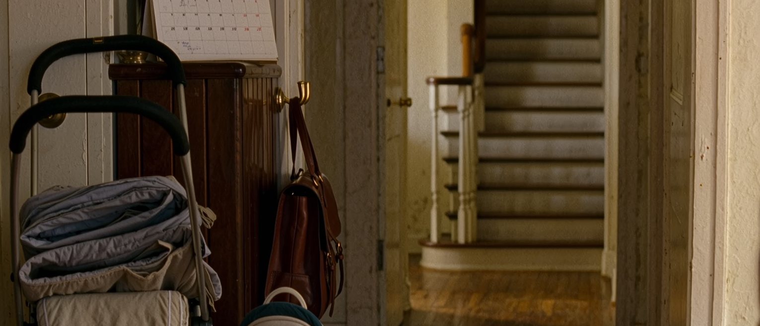 a hall seen close with a pram folded against the wall, a work satchel on the hook beside a baby carrier, a calendar with four weeks blocked in blue on the console, the stairs soft behind