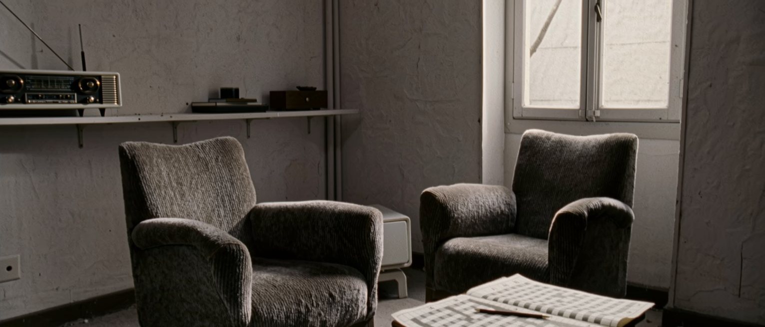 a sitting room seen close with two armchairs turned toward one another, a low table between them holding a half-finished crossword and a pencil, a radio on the shelf behind, a tall window soft beyond