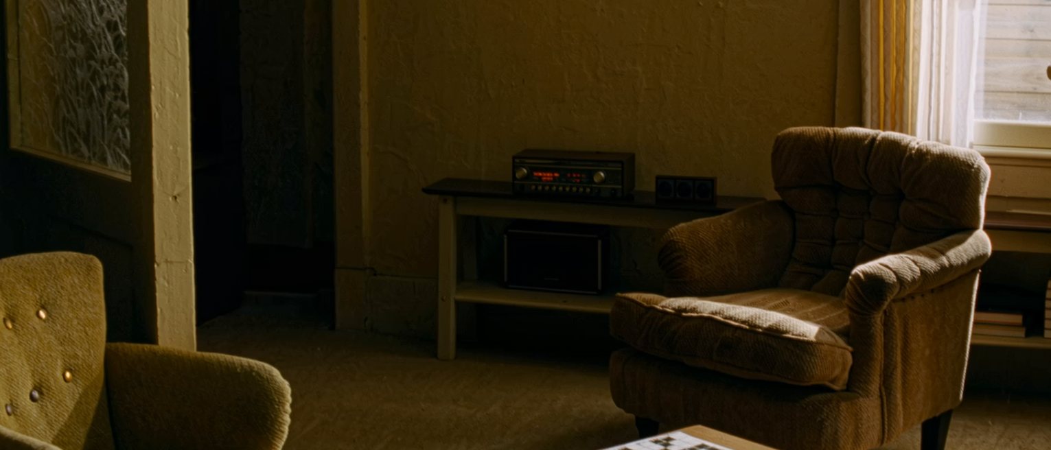 a sitting room seen close with two armchairs turned toward one another, a low table between them holding a half-finished crossword and a pencil, a radio on the shelf behind, a tall window soft beyond