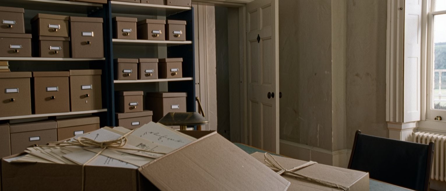 a study seen close with a shelf of labelled cardboard archive boxes, one box down on the desk with its lid off and a bundle of letters tied with string inside, a lamp and a chair, the door ajar