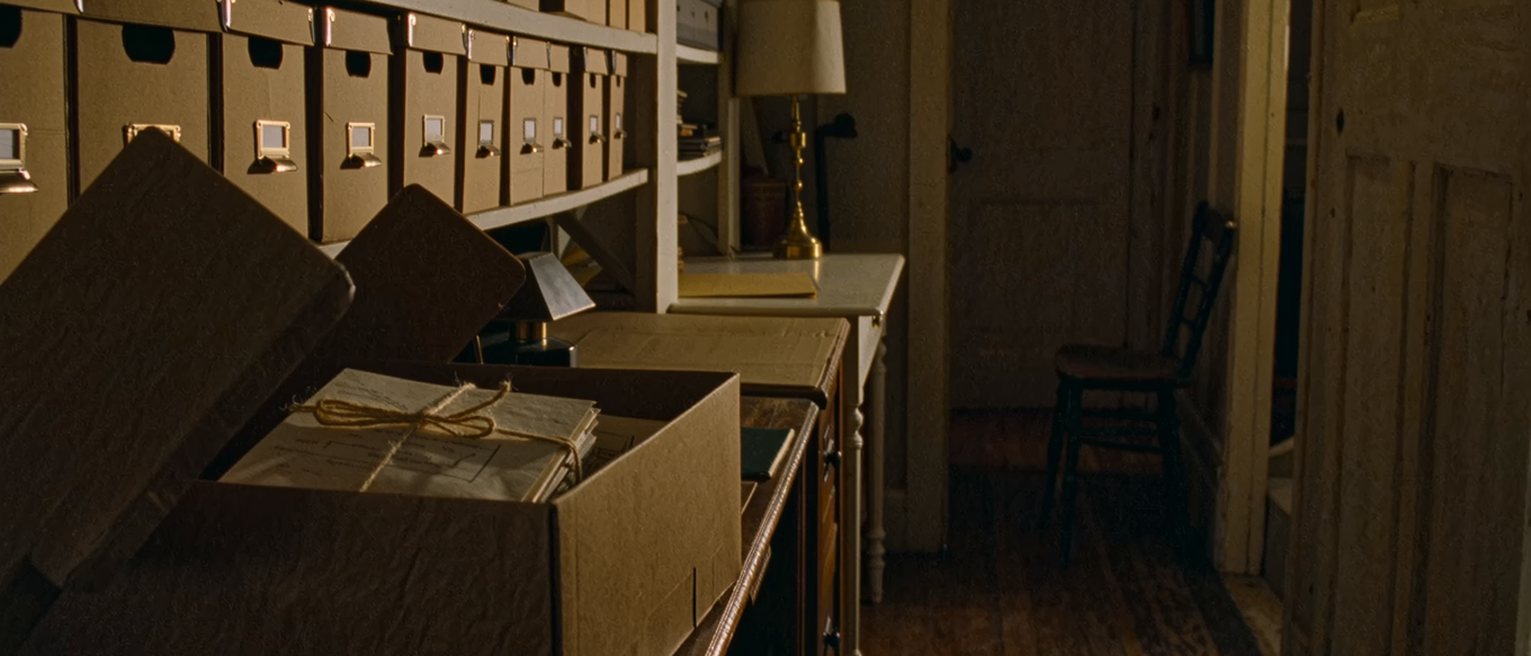 a study seen close with a shelf of labelled cardboard archive boxes, one box down on the desk with its lid off and a bundle of letters tied with string inside, a lamp and a chair, the door ajar
