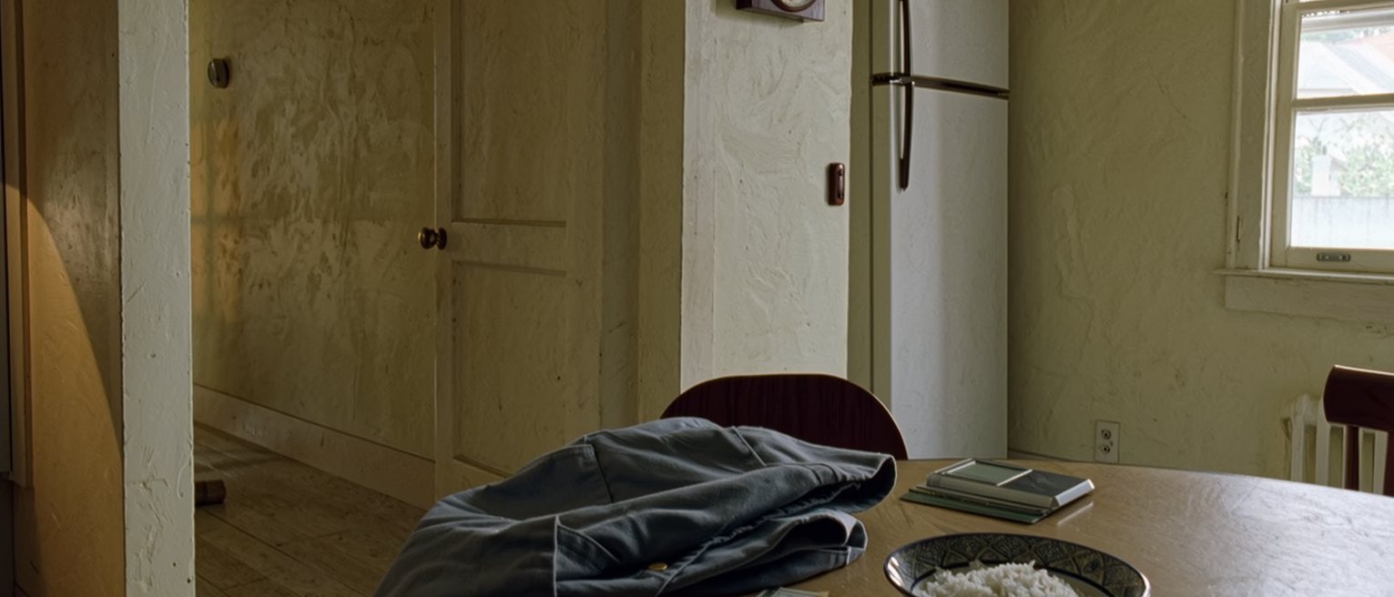 a kitchen seen close with a work uniform folded on the chair, a punch card and a payslip on the table beside a bowl of cold rice, the clock on the wall and the fridge soft behind