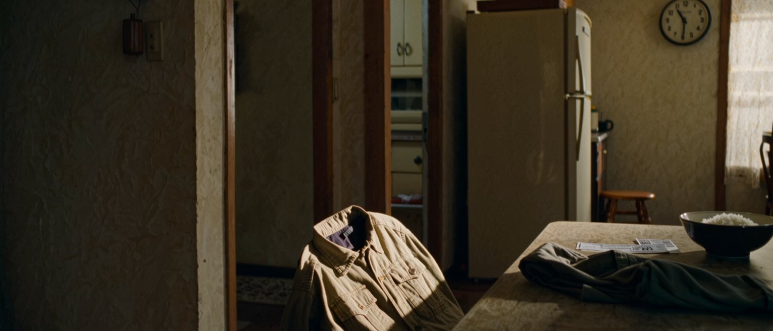 a kitchen seen close with a work uniform folded on the chair, a punch card and a payslip on the table beside a bowl of cold rice, the clock on the wall and the fridge soft behind