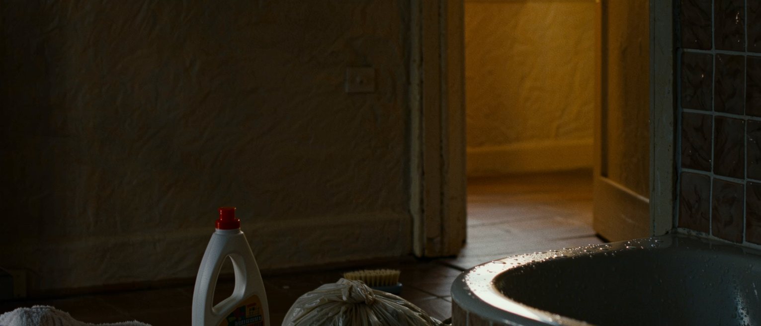 a bathroom seen close with the tub scrubbed and the tiles wet, a bottle of bleach and a brush on the floor, a folded towel and a plastic bag rolled tight, the small window soft behind