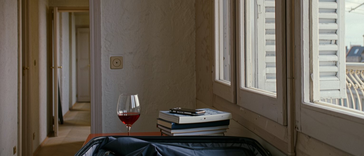 a rented flat seen close with a suitcase half unpacked, a stack of exercises on the table, a glass of wine and a phone face down, the balcony shutters with the heat outside soft behind