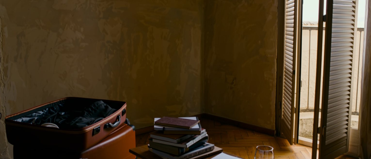 a rented flat seen close with a suitcase half unpacked, a stack of exercises on the table, a glass of wine and a phone face down, the balcony shutters with the heat outside soft behind