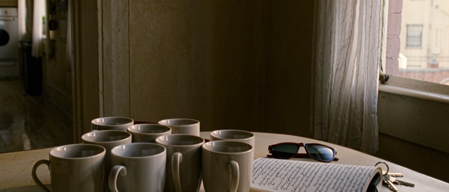 a kitchen table seen close with ten cups washed and set out to dry, a notebook open with other voices transcribed, a pair of sunglasses and a set of keys, the window onto the white city soft behind