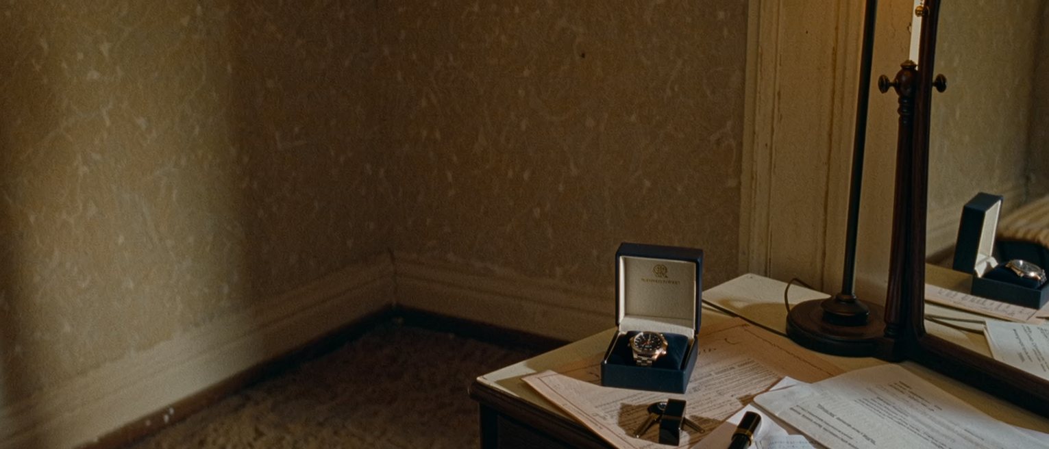 a dressing table seen close with a new wristwatch still in its box, a bank passbook open and a receipt folded beside it, a lipstick and a set of keys, the lamp close behind