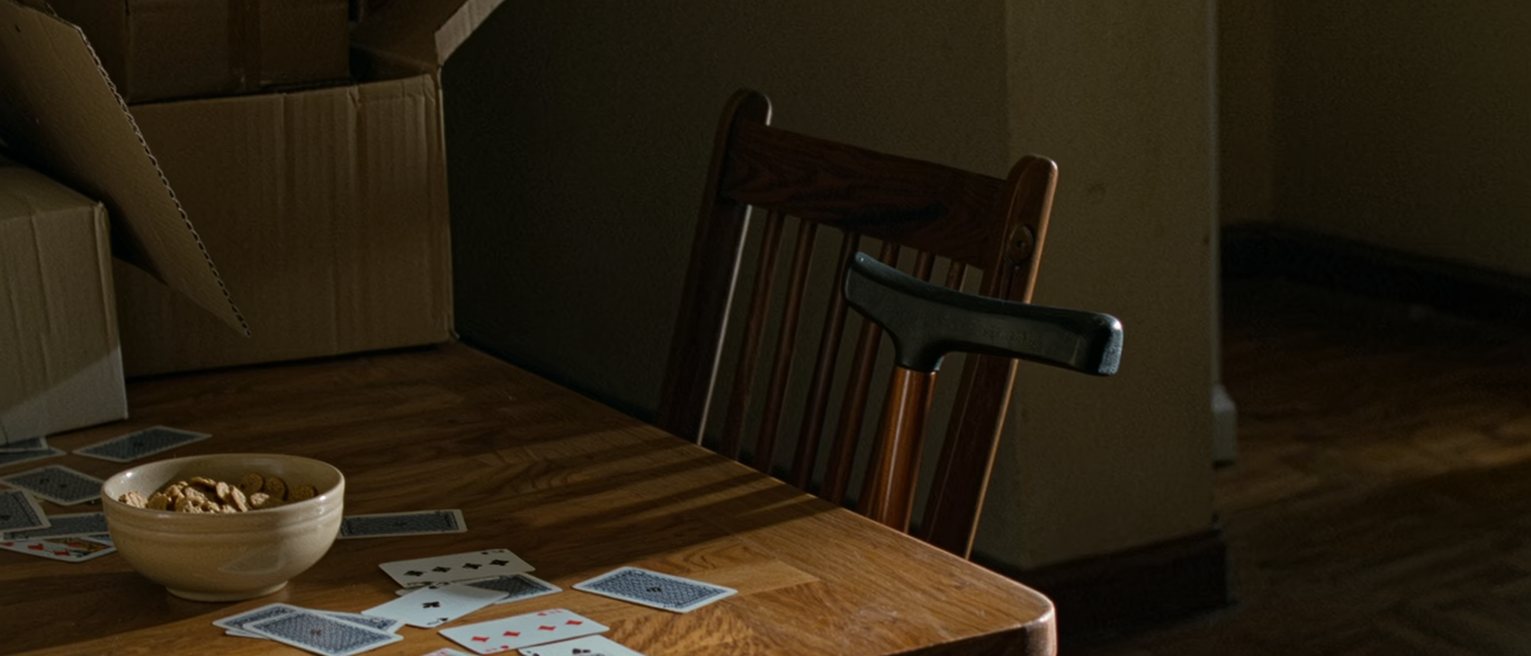 a kitchen table seen close with a bowl of cereal for one, a walking cane leaning on the chair and a pack of cards spread mid-game, the boxes of a house being emptied soft behind