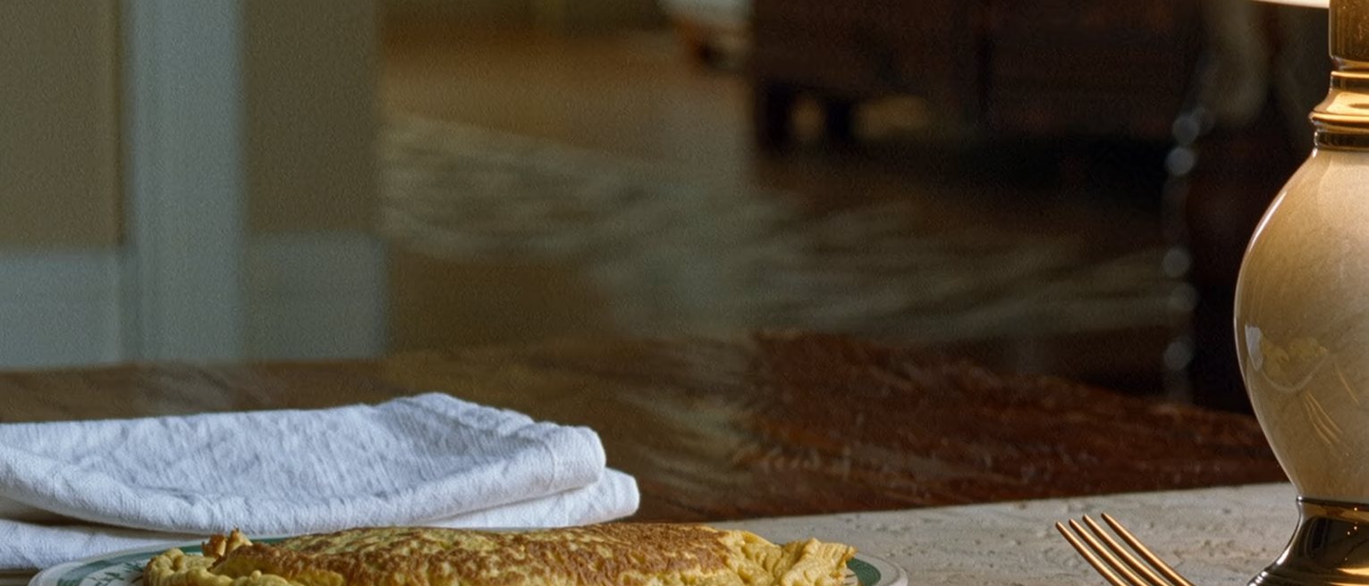 a breakfast table laid with exact precision, one place set with a folded napkin, an omelette on a plate and the fork laid at an angle that was chosen