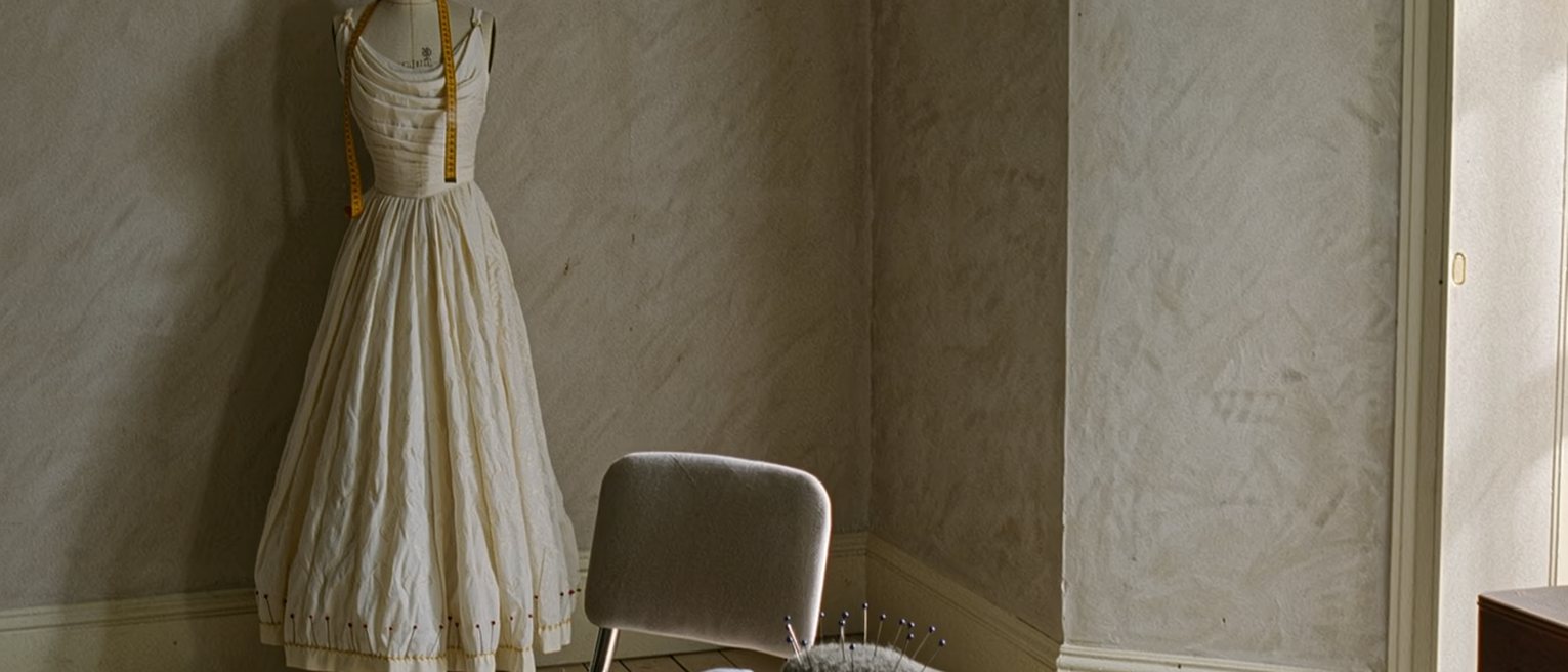 a dressmaking workroom seen close, a gown on a stand half pinned, a tape measure over the stand's shoulder, bolts of cloth and a pincushion on the table beside it and a single chair set close