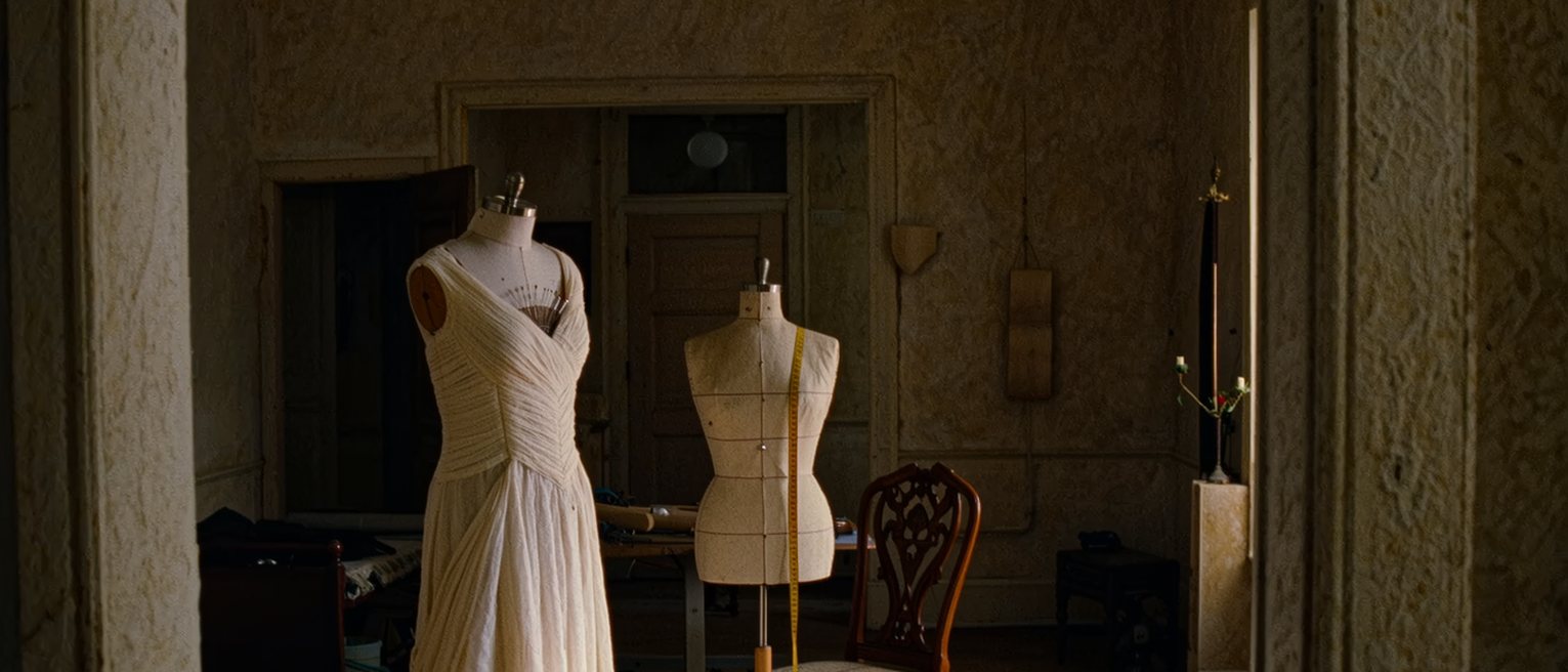 a dressmaking workroom seen close, a gown on a stand half pinned, a tape measure over the stand's shoulder, bolts of cloth and a pincushion on the table beside it and a single chair set close