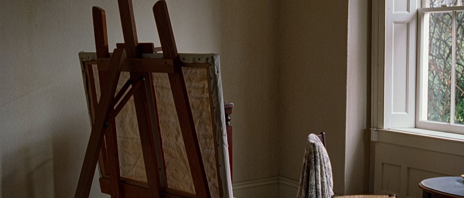 a room with an easel turned to the wall and a canvas covered by a cloth, a chair set by the window with a shawl over its back
