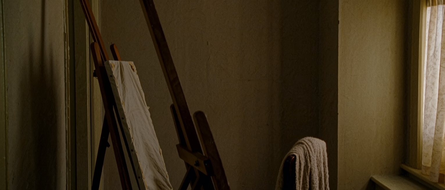 a room with an easel turned to the wall and a canvas covered by a cloth, a chair set by the window with a shawl over its back