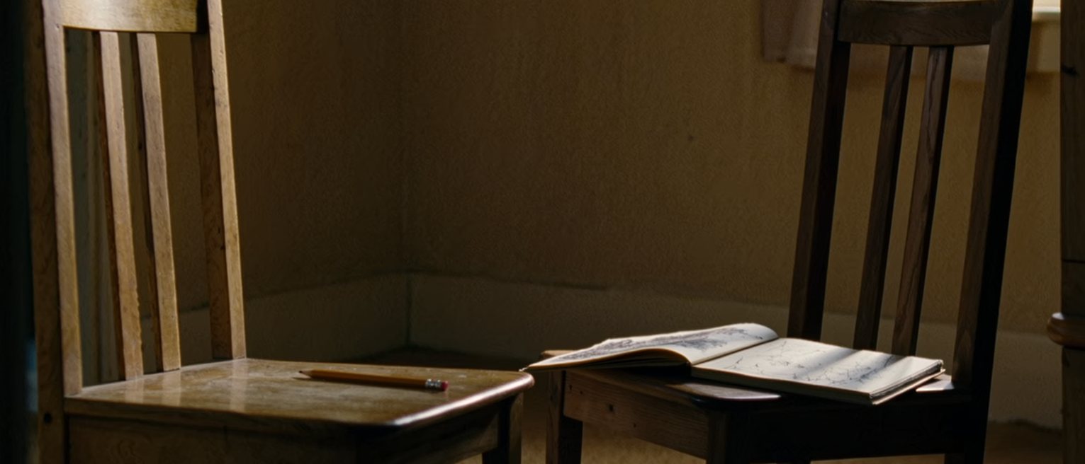 two plain wooden chairs set facing each other at a close measured distance, a sketchbook open on one of them and a pencil on the other