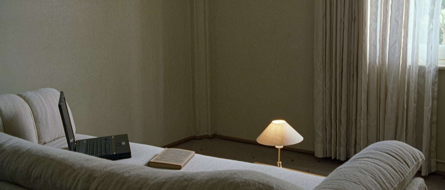a sofa seen close with two indents in its cushions at either end, a laptop open on one arm and a closed book on the other, the lamp between them lit