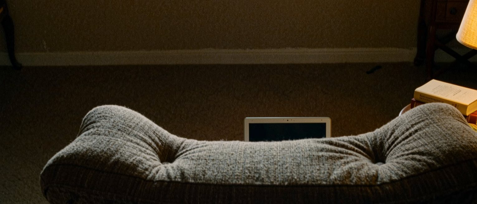 a sofa seen close with two indents in its cushions at either end, a laptop open on one arm and a closed book on the other, the lamp between them lit