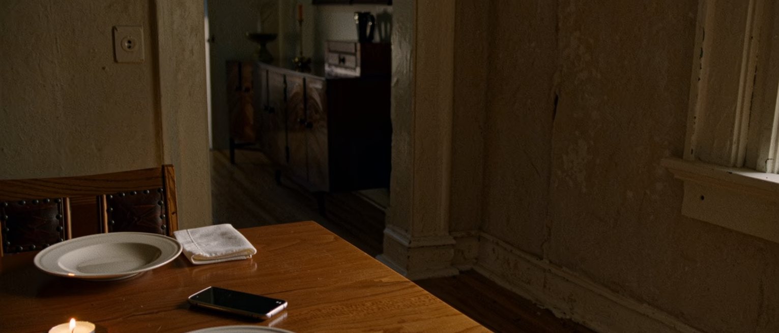 a dining table seen close after a supper for two, both plates finished and pushed together, a phone face up beside one and a napkin folded twice beside the other, a candle burned low between them and the sideboard soft behind
