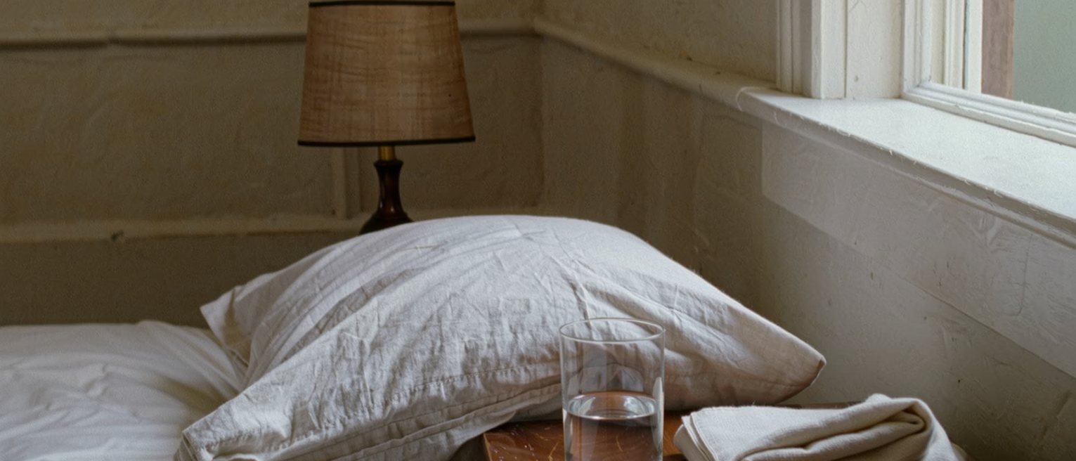 a bedside table seen close with a glass of water, a folded cloth and a small bowl of salt, the pillow dented and the lamp close behind