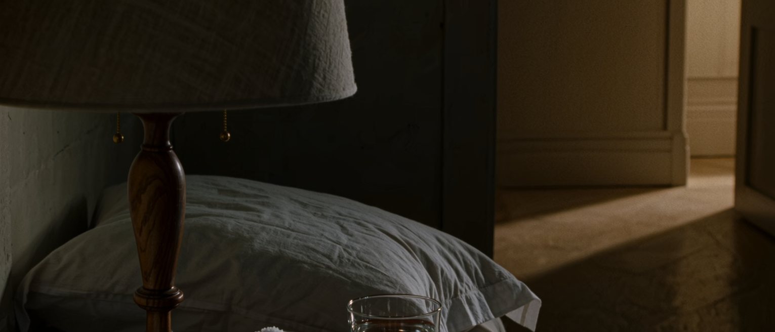 a bedside table seen close with a glass of water, a folded cloth and a small bowl of salt, the pillow dented and the lamp close behind